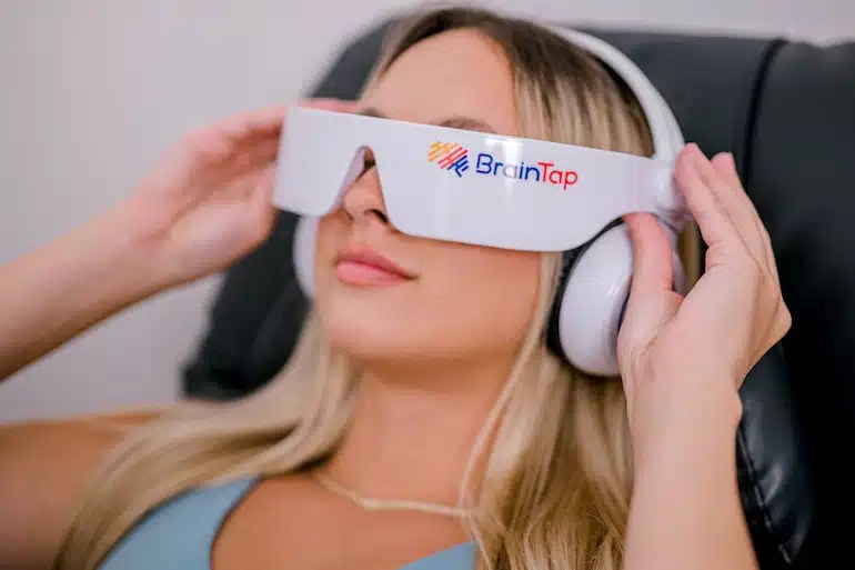 Guest reclining with eyes closed wearing a BrainTap headset and glowing light visor in a dark wellness lounge