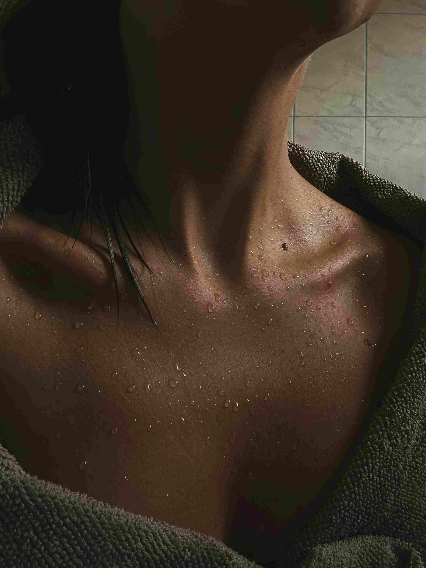 Close-up of a person's shoulder and neck covered in water droplets after a cold plunge, with a dark tiled background
